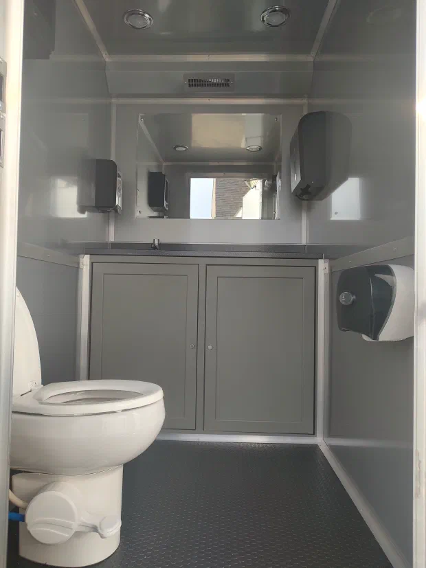 Inside a restroom trailer: a stall with a flushing toilet, a vanity and bright lighting